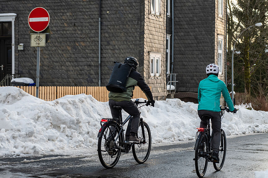 Zwei Fahrradfahrende in Winterjacken auf winterlicher Straße mit geräumtem Schnee an der Seite