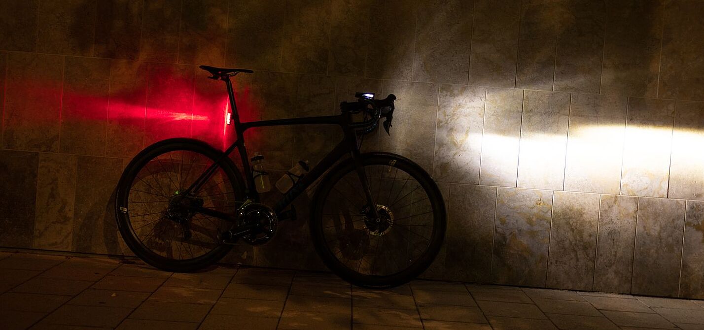 Sigma Aufmacher Ein Fahrrad steht in der Dunkelheit an einer Wand, mit am Lenker montiertem, eingeschalteten Scheinwerfer und an der Sattelstütze montierten, leuchtenden Rücklicht.