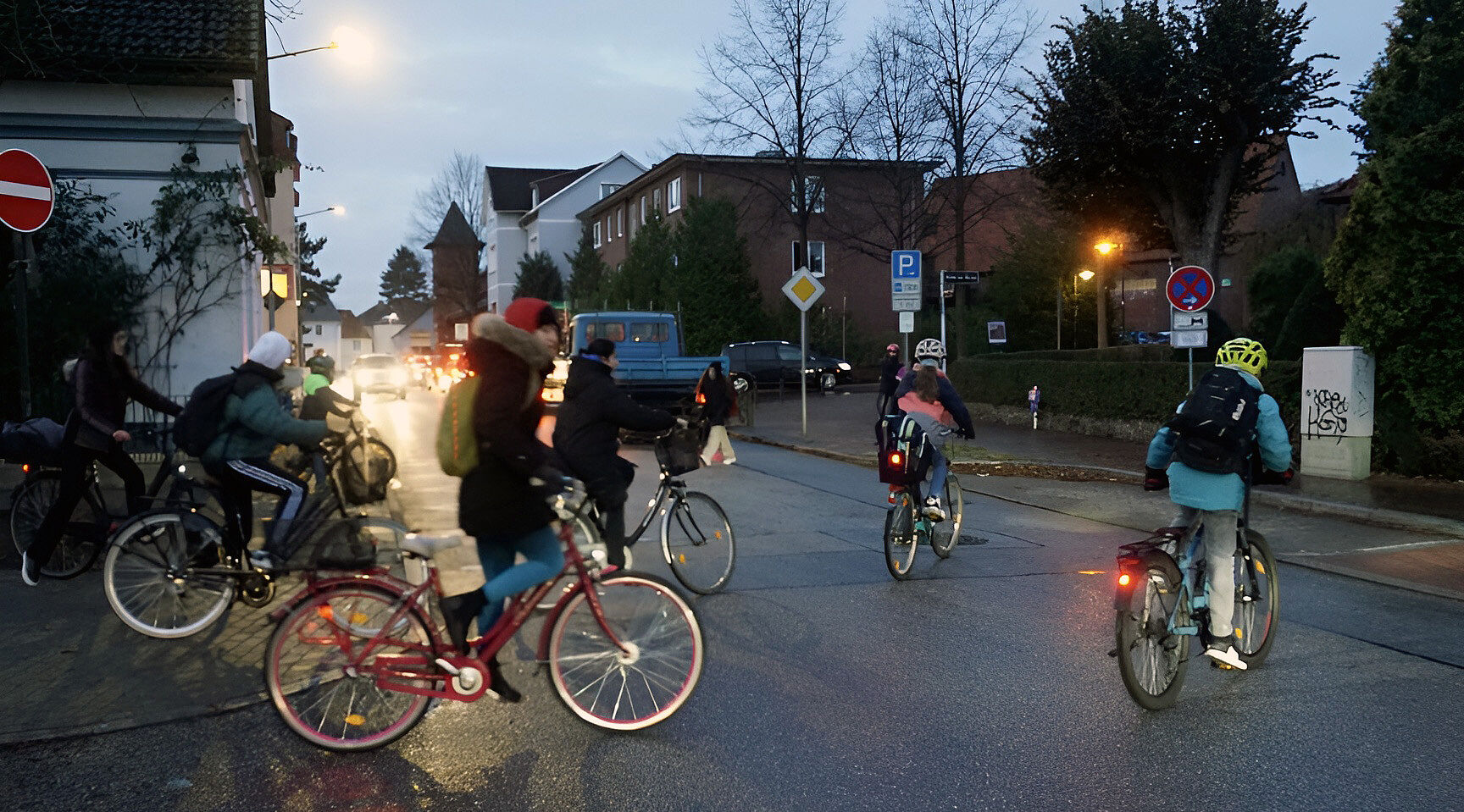 Mehrere Jugendliche fahren auf dem Rad über eine Straße, während es morgens noch dämmert. Sie tragen Winterjacken und Rucksäcke, einige schützen ihren Kopf mit einem Helm. Die Lampen an den Rädern leuchten. Im Hintergrund warten Autos mit eingeschalteten Scheinwerfern an der Straße. Straßenlaternen erhellen die Szene.