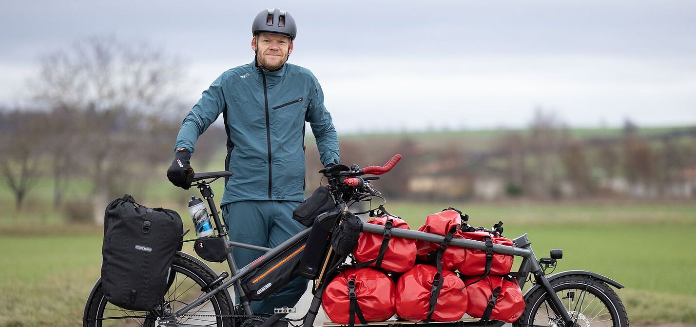 Gunnar Fehlau mit Workpacking-Lastenrad Gunnar Fehlau steht in Fahrradkleidung, vor ihm sein Lastenrad, mit dem er auf Workpacking-Tour ist.