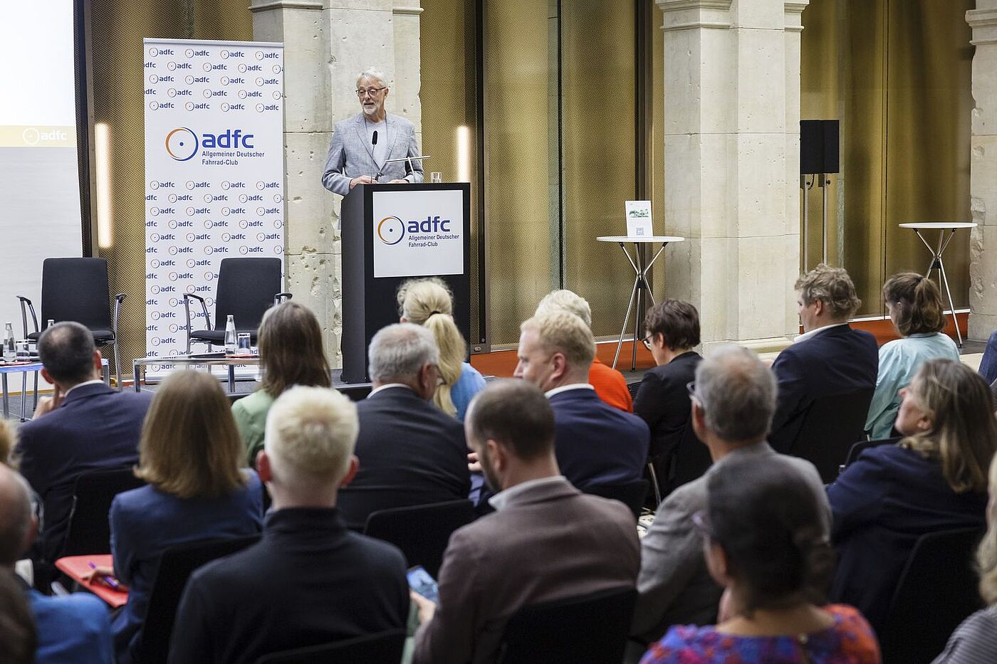ADFC-Bundesvorsitzender Frank Masurat am Pult spricht zu den Zuschauenden im Vordergrund. 