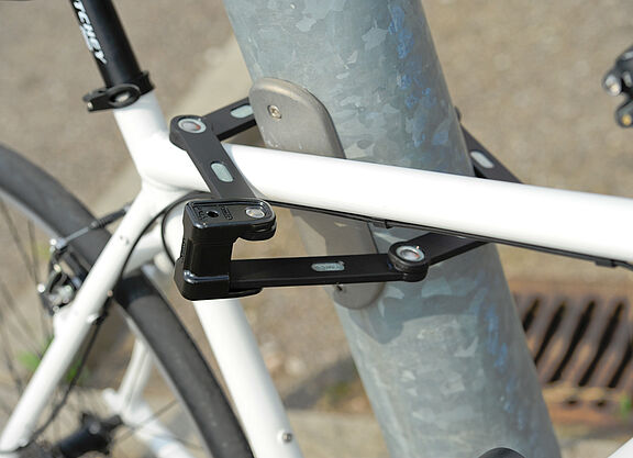 Anschließen statt nur abschließen! weißes Fahrrad ist mit einem Faltschloss an eine Metallstange angeschlossen